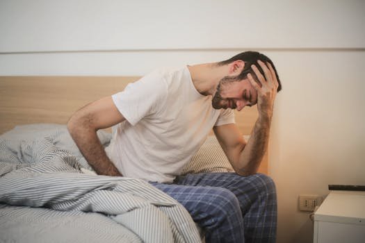 A young man in pajamas holding his head, sitting on a bed, appears to be experiencing a headache.