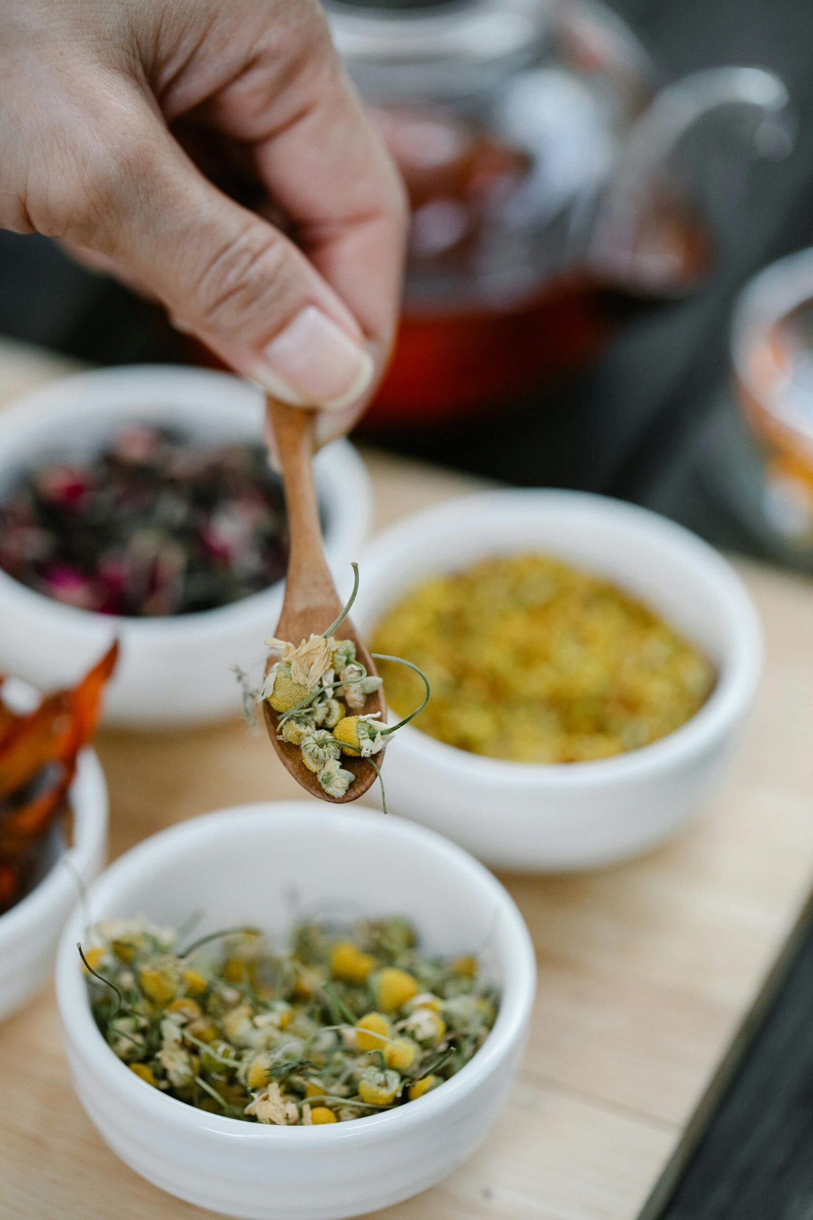 Close-up of a hand holding a wooden spoon with dried herbs and flowers in bowls.